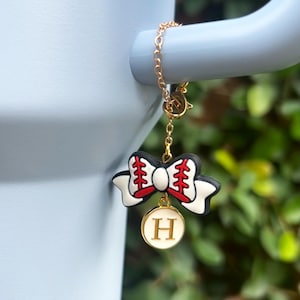 May include: A gold-colored chain with a baseball-themed charm. The charm features a white bow with black trim and red stitching, and a gold-colored circular pendant with the letter "H" in the center.