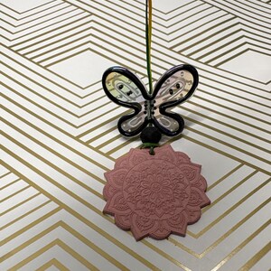 May include: A butterfly-shaped ornament with a clear body and black outline, connected to a pink, flower-shaped pendant with a detailed pattern. The ornament hangs from a rainbow-colored string.