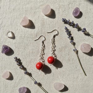 Könnte beinhalten: Silberne Kettenohrringe mit roten Perlen, präsentiert mit Lavendelzweigen und polierten Edelsteinen. Die Edelsteine umfassen Rosenquarz und Amethyst. Der Hintergrund ist ein strukturiertes Off-White.