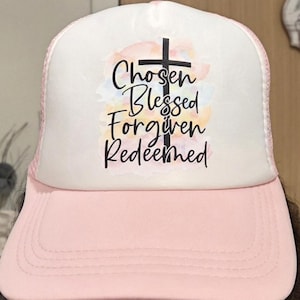 May include: A pink and white trucker hat with the words "Chosen, Blessed, Forgiven, Redeemed" in black script, with a black cross. The hat has a pink brim and a white mesh back.