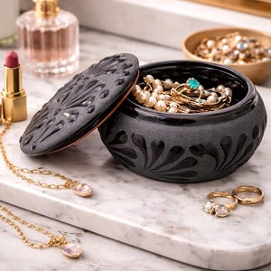 Op de afbeelding: Een zwarte sieradendoos met een decoratief deksel, gevuld met parels en gouden ringen. Gouden kettingen met roze edelstenen en gouden ringen liggen op een marmeren oppervlak. Een parfumfles en lippenstift zijn ook zichtbaar.