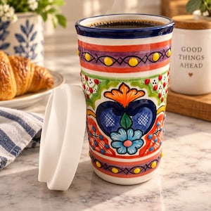 May include: A tall ceramic travel mug featuring a vibrant, hand-painted floral and geometric pattern in shades of red, blue, green, orange, and yellow. The mug has a dark blue rim, a white silicone lid, and raised dot details throughout the design.