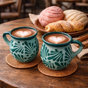 May include: Two teal mugs with a floral design and heart-shaped latte art sit on woven coasters. A wooden plate holds Mexican sweet bread, including conchas. The scene is set on a wooden table, suggesting a cafe setting.