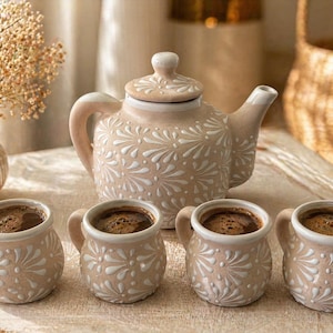 Puede incluir: Un juego de té de cerámica beige con una tetera y cuatro tazas pequeñas, todas llenas de café. El juego presenta un patrón floral blanco. La tetera tiene tapa y asa. Las tazas tienen asas y están dispuestas en fila.