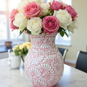 May include: A decorative pink and white vase filled with pink and white roses. The vase features a detailed floral design. The arrangement is on a marble table, creating a bright and elegant display.