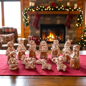 Könnte beinhalten: Eine Weihnachtskrippe mit Terrakottafiguren und Hunden auf einer roten Tischdecke. Die Figuren sind mit weißen Akzenten bemalt. Im Hintergrund ein Kamin mit Feuer und Weihnachtsdekorationen.