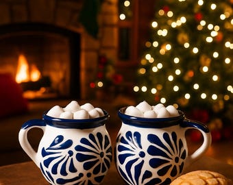 Op de afbeelding: Twee wit en blauwe keramische mokken gevuld met marshmallows staan op een houten tafel, naast een zoet broodje. De mokken hebben een bloemenontwerp. Een kerstboom en een open haard zijn op de achtergrond.