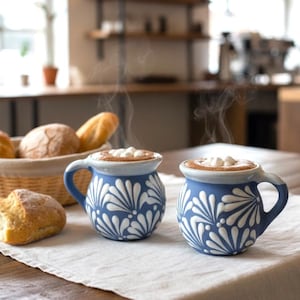 May include: Two blue and white ceramic mugs with handles, filled with a hot beverage and topped with marshmallows. The mugs have a floral design. A basket of bread and a loaf of bread are in the background.