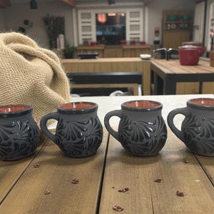 Może przedstawiać: Cztery szare ceramiczne kubki z ciemnym kwiatowym wzorem i brązowym wnętrzem ustawione na drewnianej powierzchni. Każdy kubek ma uchwyt i zaokrąglony kształt. Kubki są częścią zestawu naczyń kuchennych.