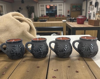 May include: Four gray ceramic mugs with a dark floral design and a brown interior are arranged on a wooden surface. Each mug has a handle and a rounded shape. The mugs are part of a kitchenware set.