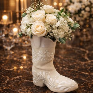 Può includere: Un vaso a forma di stivale da cowboy in ceramica color crema, ornato da un motivo floreale in rilievo, contenente un bouquet di rose bianche e verde. Il vaso è posto su una superficie di marmo marrone.