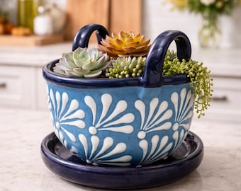 May include: A blue ceramic basket planter with white floral designs, filled with various succulents. The planter has two arched handles and sits on a matching saucer. The arrangement is on a countertop.