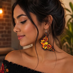 Op de afbeelding: Een kralen vlinder oorbellen in tinten oranje, geel en zwart. De oorbel wordt op het oor gedragen. De vrouw draagt een zwarte off-the-shoulder top met bloemenborduurwerk.