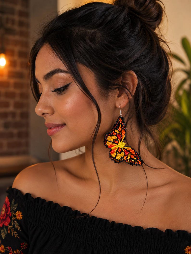 Op de afbeelding: Een kralen vlinder oorbellen in tinten oranje, geel en zwart. De oorbel wordt op het oor gedragen. De vrouw draagt een zwarte off-the-shoulder top met bloemenborduurwerk.