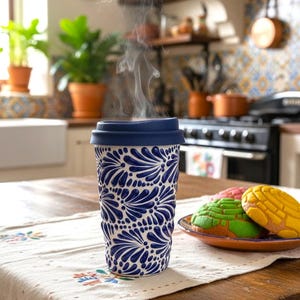 May include: A ceramic travel mug with a blue floral pattern and a blue lid. Steam rises from the mug, suggesting a hot drink. A plate of colourful pastries sits nearby on a wooden table.