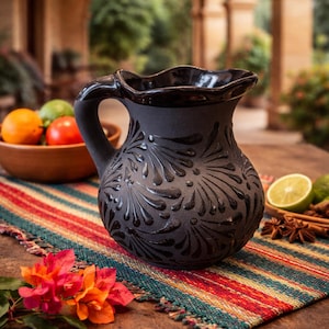May include: A black ceramic pitcher with a decorative floral pattern. The pitcher has a handle and a ruffled rim. It sits on a colorful striped textile, with a bowl of fruit and spices nearby. The scene is set outdoors.