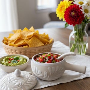 Peut inclure: Une table garnie de chips tortilla, de guacamole et de salsa dans un bol décoratif avec une poignée. Un vase de fleurs colorées est également présent. Le bol de salsa a un couvercle.