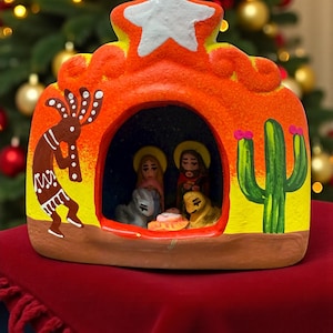 Könnte beinhalten: Eine handbemalte Krippe aus Keramik in leuchtenden Farben Orange, Gelb und Grün. Die Szene zeigt einen Stern, eine Kokopelli-Figur, einen Kaktus und die Heilige Familie. Die Basis befindet sich auf einer roten Oberfläche, mit einem Weihnachtsbaum im Hintergrund.