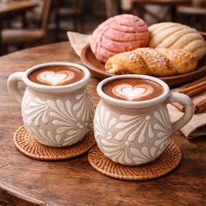 May include: Two mugs of hot chocolate with heart-shaped latte art sit on woven coasters. A wooden plate holds sweet bread, including a pink concha. The scene is set on a wooden table, suggesting a cafe or bakery.