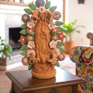 May include: A terracotta statue of a religious figure, adorned with colorful flowers and leaves. The statue is placed on a dark wooden side table. The background includes a fireplace and a patterned armchair.