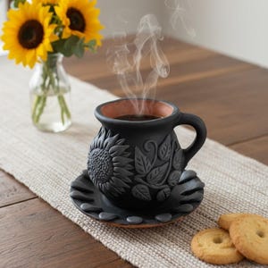May include: A black ceramic coffee cup and saucer set with a sunflower design. The cup contains a dark liquid, with steam rising. The set is on a table with a vase of sunflowers and a few cookies.