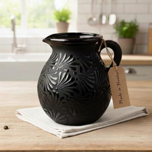 Może przedstawiać: Czarny ceramiczny dzban z dekoracyjnym wzorem kwiatowym. Dzbanek ma uchwyt i dzióbek do nalewania. Stoi na białej serwetce na drewnianej powierzchni. Do uchwytu przymocowana jest etykieta z napisem "Made in Mexico".