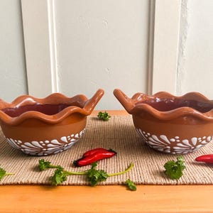 Może przedstawiać: Dwie ceramiczne miski w kolorze terakoty z falistymi krawędziami i uchwytami. Miski mają biały kwiatowy wzór wokół podstawy. Czerwone papryczki chili i zielone zioła ułożone są wokół misek na tkanej macie.
