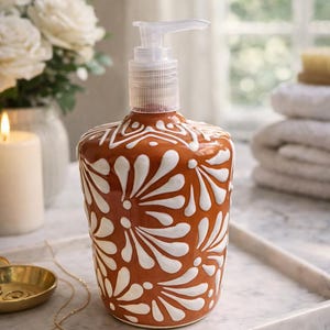 May include: A terracotta-coloured ceramic soap dispenser with a white floral design and a clear pump. The dispenser is on a marble tray. The background includes a candle, flowers, and towels.