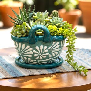 May include: A teal ceramic planter filled with various succulents. The planter has a handle and a white floral design. The arrangement sits on a matching saucer and a striped blue and white placemat, on a wooden table.