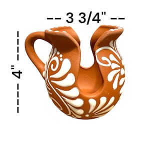 Op de afbeelding: Een terracotta keramische kan, ongeveer 10 cm hoog en 9,5 cm breed, met een handvat en een unieke U-vormige opening. De kan is versierd met witte bloemen- en wervelpatronen, wat de rustieke uitstraling versterkt.