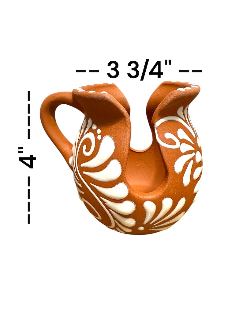 Op de afbeelding: Een terracotta keramische kan, ongeveer 10 cm hoog en 9,5 cm breed, met een handvat en een unieke U-vormige opening. De kan is versierd met witte bloemen- en wervelpatronen, wat de rustieke uitstraling versterkt.