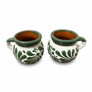 May include: Two handmade ceramic mugs. The mugs are white with green floral designs and green rims. The interior of the mugs is a warm brown color. Each mug has a small handle. The mugs are likely used for drinking beverages.