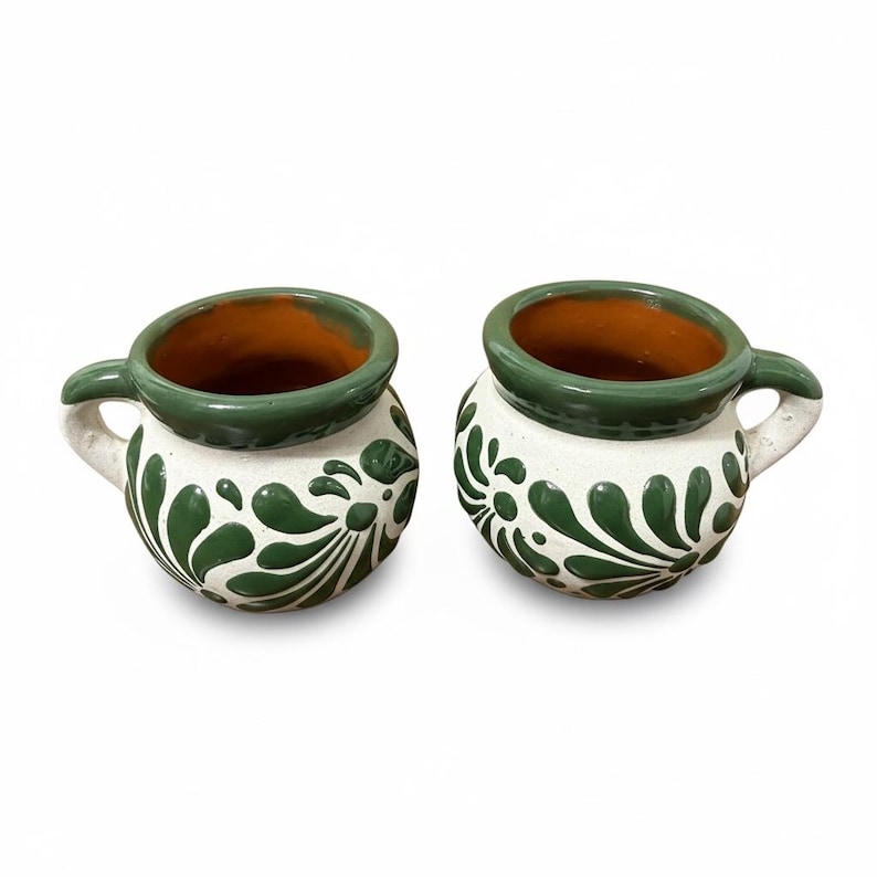 May include: Two handmade ceramic mugs. The mugs are white with green floral designs and green rims. The interior of the mugs is a warm brown color. Each mug has a small handle. The mugs are likely used for drinking beverages.