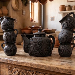 May include: A black ceramic tea set, including a teapot and stacked teacups, adorned with a detailed floral design. The set is presented on a rustic wooden table, set against a warm kitchen background.
