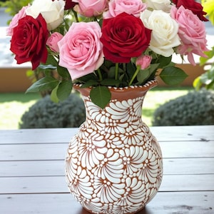 Puede incluir: Un ramo de rosas rojas, rosas y blancas en un jarrón decorativo. El jarrón es marrón con un diseño floral blanco. Las flores son frescas y vibrantes, con hojas verdes. El arreglo está sobre una mesa de madera.