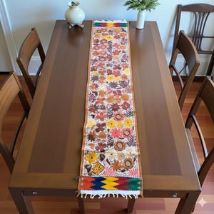 Puede incluir: Un camino de mesa rectangular con estampado floral y borde marrón, colocado sobre una mesa de madera. El camino de mesa presenta una variedad de flores coloridas en tonos naranja, amarillo y marrón. La mesa está rodeada de sillas de madera y se ve un jarrón.