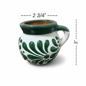 May include: A handcrafted ceramic mug, 3 inches tall and 2.75 inches wide, with a white base and green floral design. The rim and handle are green, and the interior is orange. The mug has a rounded shape and a small handle.