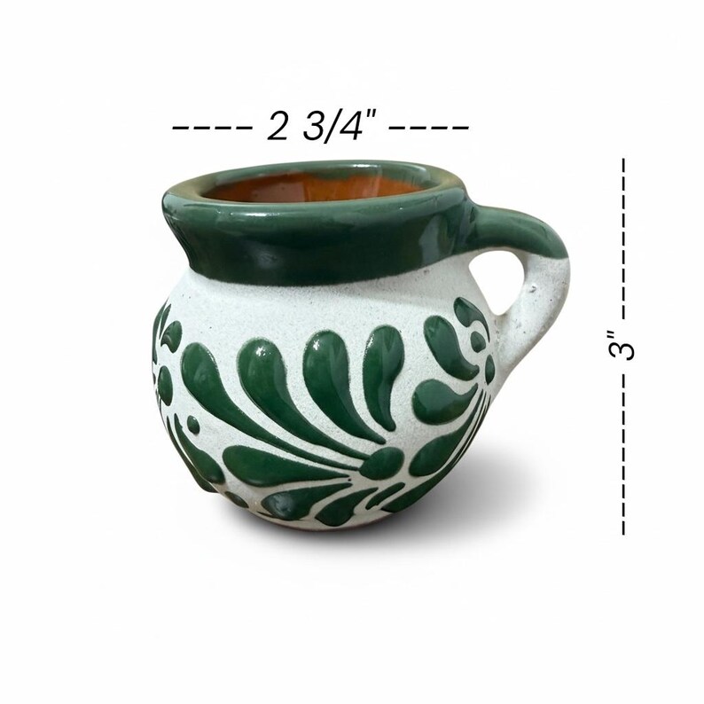May include: A handcrafted ceramic mug, 3 inches tall and 2.75 inches wide, with a white base and green floral design. The rim and handle are green, and the interior is orange. The mug has a rounded shape and a small handle.