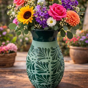 May include: A colourful floral display in a green vase. The bouquet features sunflowers, roses, and other bright flowers. The vase has a textured, embossed pattern. The arrangement is on a wooden surface.