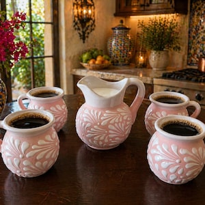 May include: A collection of pink ceramic coffee mugs and a creamer with white floral designs. The mugs are filled with dark coffee and arranged on a wooden table. A vase of flowers is in the background.