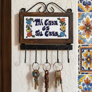 Könnte beinhalten: Ein Wand-Schlüsselhalter mit einem dunklen Holzrahmen und einem dekorativen Kacheleinsatz. Auf der Kachel steht "Mi Casa es Su Casa" und ist mit floralen Akzenten verziert. Darunter befinden sich fünf schwarze Haken mit Schlüsseln.
