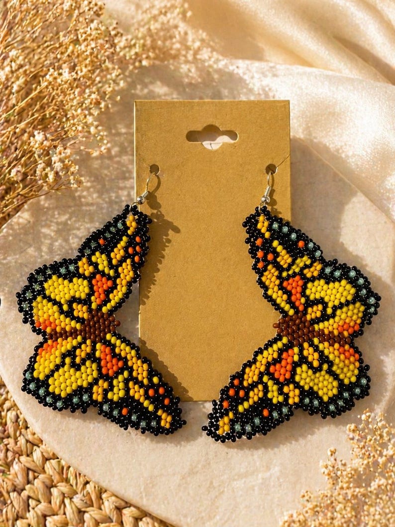 Op de afbeelding: Een paar geborduurde vlinder oorbellen. De oorbellen hebben gele, oranje en zwarte kralen, met een bruin midden. De vlindervleugels zijn gedetailleerd met een verscheidenheid aan kralenkleuren. De oorbellen worden gepresenteerd op een bruine kaart.