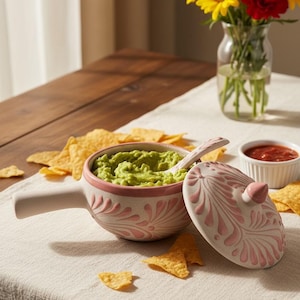 May include: A ceramic serving bowl filled with guacamole, a spoon, and a matching lid. The bowl and lid are white with pink floral designs. Tortilla chips and a small bowl of salsa are also present, with a vase of yellow and red flowers.