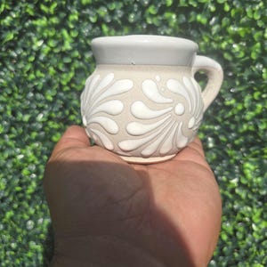 May include: A small, beige ceramic mug with a white floral design and a gray rim and handle. The mug is held in a hand against a green background.