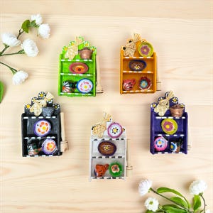 Può includere: Cinque mensole decorative in miniatura, colorate, con piccoli piatti e ciotole. Ogni mensola è di un colore diverso: verde, arancione, nero, bianco e viola. Le mensole sono in legno e hanno un aspetto rustico e artigianale.