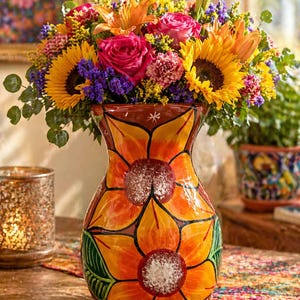 May include: A vibrant floral arrangement in a hand-painted ceramic vase. The vase features large orange and yellow flower designs. The bouquet includes sunflowers, roses, and lilies in shades of yellow, orange, pink, and purple. The vase sits on a colorful patterned table runner.