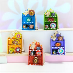 Può includere: Cinque piccole mensole da cucina in legno colorato, ognuna dipinta con un colore diverso: blu, verde, giallo, rosso e viola. Ogni mensola contiene piatti, tazze e un mattarello in miniatura. Le mensole sono decorate con fiocchi di paglia.