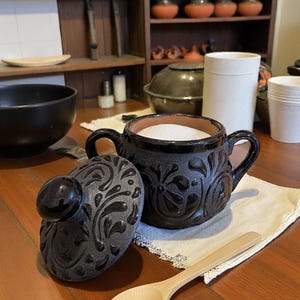 May include: A dark brown ceramic sugar bowl with a lid, filled with white sugar, and a wooden spoon. The bowl has two handles and floral relief patterns. The lid is placed next to the bowl on a white cloth. A wooden shelf with mugs is in the background.