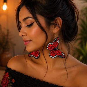 Op de afbeelding: Rode en zwarte kralen vlinder oorbellen. De oorbellen zijn felrood met zwarte details, gevormd als vlinders. De oorbellen worden gedragen met een zwarte off-the-shoulder top met rode bloemenborduurwerk.