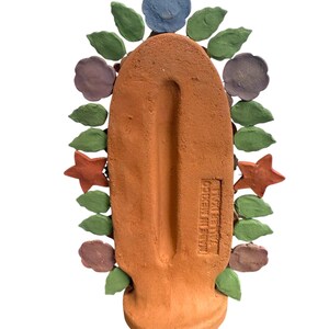 May include: A terracotta-colored, handcrafted decorative piece with a central, elongated shape. It is adorned with colorful, stylized flowers, leaves, and stars in shades of blue, green, purple, and orange. The text "MADE IN MEXICO" is visible.